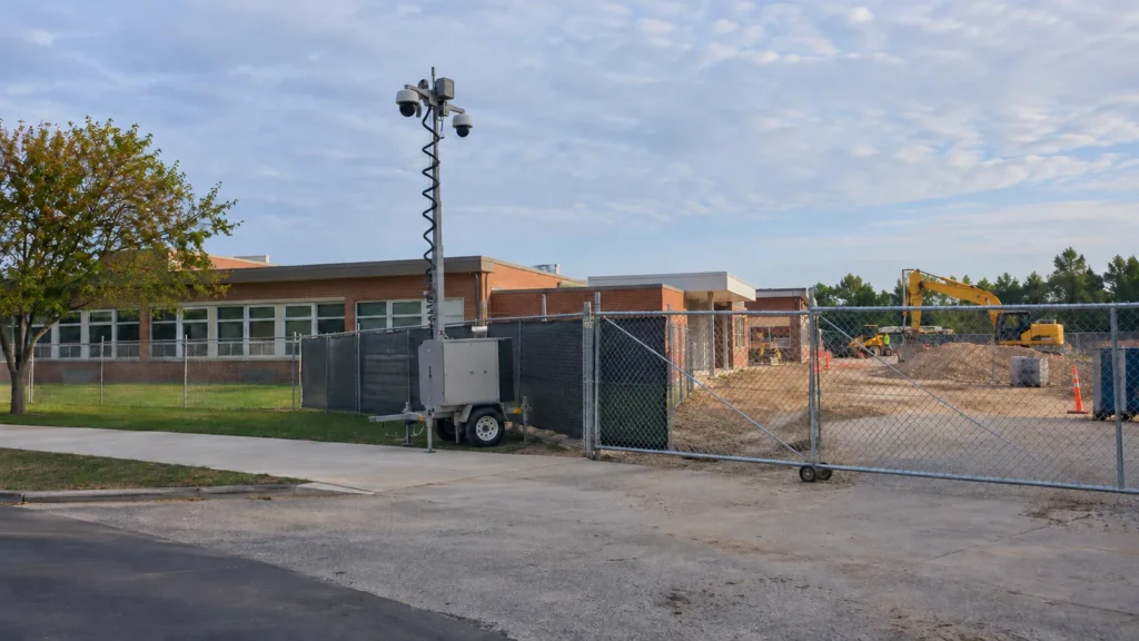 Fenced construction zone beside a school building with a surveillance trailer at the site entrance